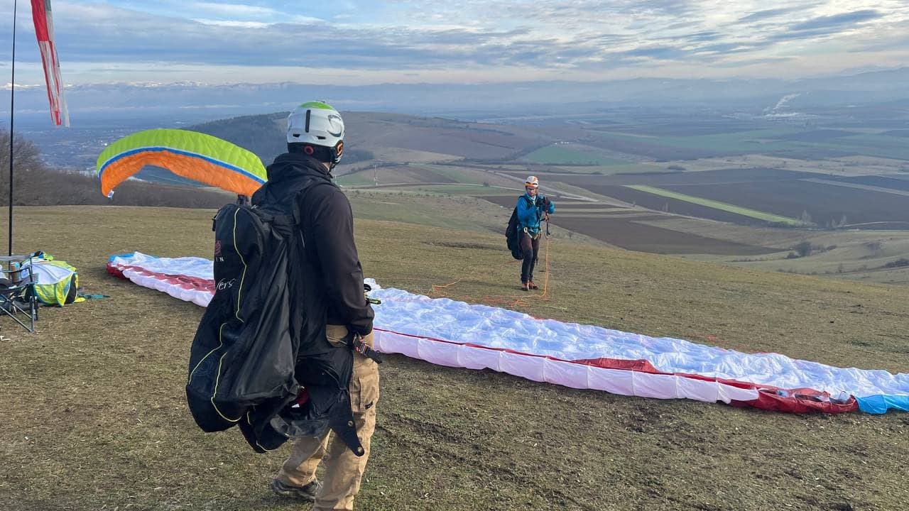 Pilot cu rucsacul în spate lângă o aripă întinsă pe deal, în timp ce altul decolează